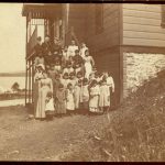 Staff and children standing posed for a portrait on the steps coming off the side of Crosby Girls' Home, mountains and water in the background.