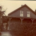 Mission house at port Simpson with two individuals standing on the verandah, shrubs in front of the house and part of the path leading up to the house visible.