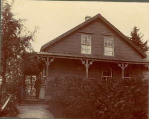 Mission house at port Simpson with two individuals standing on the verandah, shrubs in front of the house and part of the path leading up to the house visible.