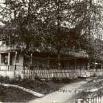 Hospital with large tree in front, surrounded by picket fence and path leading up to it.