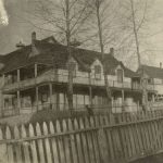 Hospital with three trees in front of it, partial view of picket fence in the foreground.
