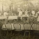 Children setting tables with white tablecloths, outdoors, trees in the background.