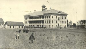Children planting in a field with the Portage la Prairie Residential School behind them