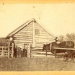 People posed in front of wood cabin, ox cart to the side
