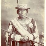 Saulteaux person posed with wooden staff, caption reads 