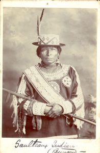 Saulteaux person posed with wooden staff, caption reads 