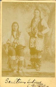 Two Saulteaux people, one seated one standing for portrait indoors with a backdrop of a tree. Caption in cursive writing at bottom of the image reads 