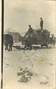 Rev. Charles Hopkins and another person on freight sleigh, lead by horses in the winter. School in the background.