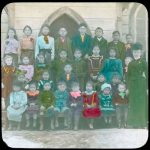 Large group of children seated and standing in rows in front of a building. Image has had colour added by paint.