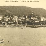 View of Lax Kw'alaams, seen from the water. Buildings clustered at the shoreline with heavy forest on the hills in the background. Type text on top of image reads 
