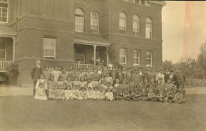 Children and staff gathered together, some are standing in the back of group, with some kneeling, and the front row seated on grass. The institution building is seen behind the group.
