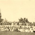 Children and youth outside in a group. The front row is seated on the grass, with a row kneeling, and two rows of standing people, tallest in the final row. Staff are part of the final row.