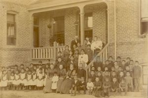 Children gathered on and around a porch. Those on the left of the porch are wearing dresses and aprons, those on the right of the porch are wearing pants and shirts. The porch in the centre has staff and some children. A large dog is laying on the ground looking at the camera in the right foreground.