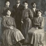 A group of six people, two are seated, with the remaining four standing between and behind the two seated people.