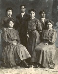 A group of six people, two are seated, with the remaining four standing between and behind the two seated people.