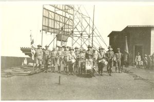 Scouts and band standing in uniforms on roof, entrance and scaffolding behind them, band holding drums