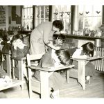 Children writing at desks, staff member is leaning over a child and writing