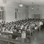 Boys seated in multiple rows of assembly hall, some are standing at side