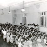 Girls seated in multiple rows of assembly hall, some are standing at side