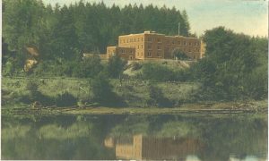 View of Alberni Indian Residential School building from water, building surrounded by forest, hand coloured