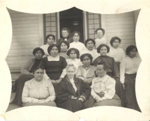 Group of 16 people seated and standing on the front steps of Kitimaat Residential School.