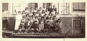 Large group of children and staff seated and standing posed for a portrait on the steps of Kitimaat Residential School.
