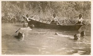 Children swimming and rowing in a boat, close to shore, line of trees in the background.