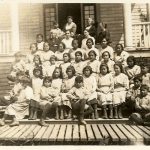 Large group of children seated and standing on the front steps of Kitimaat Residential School with staff behind them.