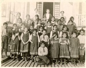 Children standing in rows posed with staff behind them, on the steps of Kitimaat Residential School.