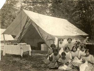 Group of youth with staff sitting and sewing beside a large canvas tent, with a table in front of it, trees in the background.