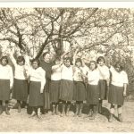 Youth standing in a line outdoors and waving at the camera, Miss Currie at centre. Trees in the background.