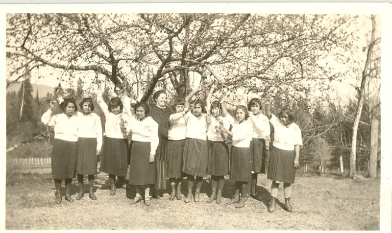 Youth standing in a line outdoors and waving at the camera, Miss Currie at centre. Trees in the background.