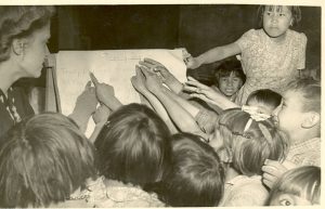 Children surrounding and pointing at lesson on easel, staff member to the side