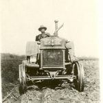Person wearing hat driving plough in field