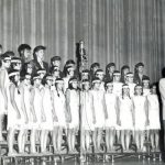 Glee Club performing, standing on three tiers on a stage in clothing for the performance, adult in front leading