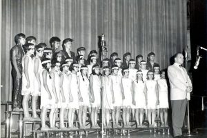 Glee Club performing, standing on three tiers on a stage in clothing for the performance, adult in front leading