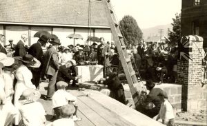 Large group assembled outdoors in the sun to watch laying of the cornerstone, building in the background and ladder leaning against building.