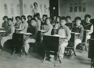 Children sitting in their desks all facing camera with teacher in the centre back of the classroom.