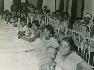 Two rows of children in their beds under the blankets, one holds a doll.