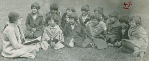 Teacher holds a book while sitting on the grass with group of students.