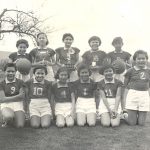 Youth in soccer uniforms, seated and standing with trophies and basketballs., outdoors.