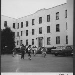Group of children running across the road in front of Alberni Residential School, some cars parked in front of the building.