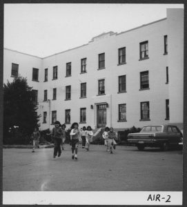 Group of children running across the road in front of Alberni Residential School, some cars parked in front of the building.