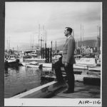 Rev. F. E. (Ed) Kempling standing on a dock looking out at a harbour filled with boats.