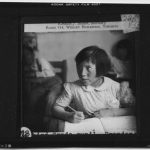 Child seated at a desk, pencil in hand writing in a book, scan of a lantern slide.