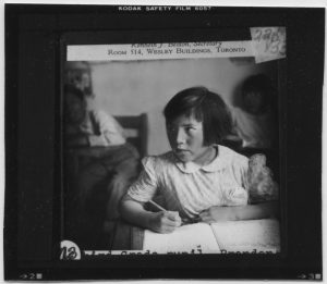 Child seated at a desk, pencil in hand writing in a book, scan of a lantern slide.