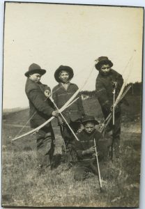 Four children, three standing, with one kneeling in foreground, holding archery equipment.