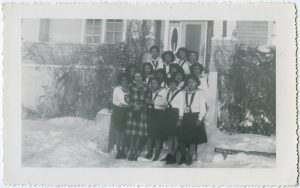 Group of youth standing posed for a photograph in Canadian Girls in Training Uniforms with dietician Allison McLean in front of Round Lake Residential School in winter.