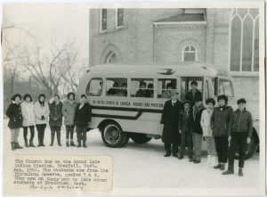 Group of students and Rev. Earle Stotesbuty posed outside of a school bus, building in the background, in winter. Label affixed to the bottom reads what image title says.