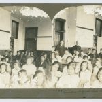 Large group of youth seated facing forwards with staff members standing at the back of the room. Image is a stereograph.